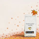 A bag of Sumiyaki Coffee stands on a surface covered with scattered autumn leaves against a pale cream background. The bag is white with black text reading 'SUMIYAKI coffee' and a small illustration of a hand holding a cup.