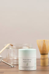 Matcha powder container on a wooden surface with tea-making tools in the background