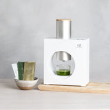 White bottle with silver cap on a wooden surface, with a glass of green liquid and small containers nearby.